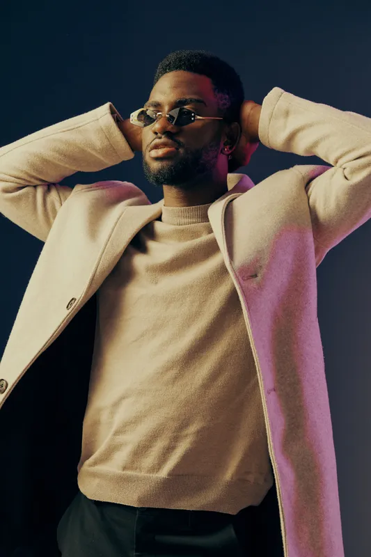 Stylish Black man in an elegant coat and sunglasses, showcasing confidence against a dark background with vibrant lighting, perfect for fashion and lifestyle related themes