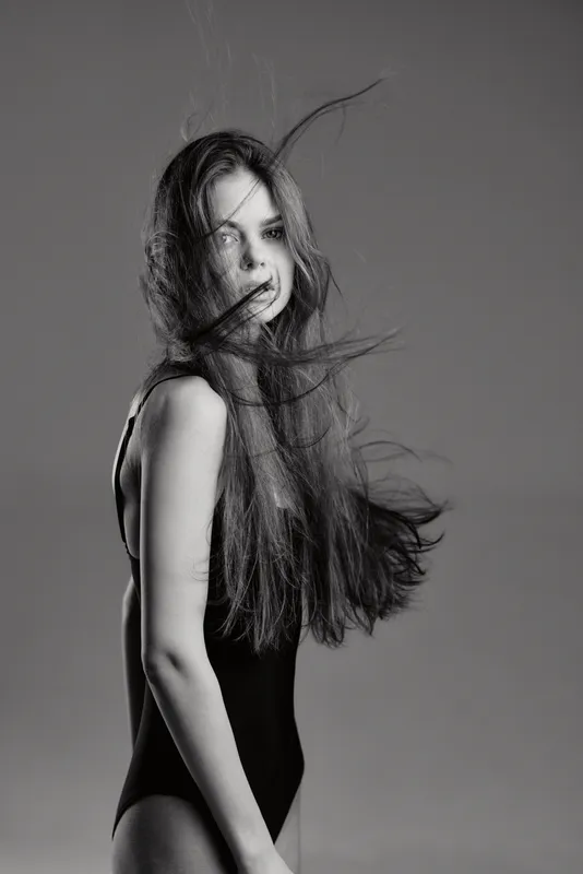 Black and white portrait of a young woman with long hair blowing, wearing a sleeveless top, expressing calm and elegance.