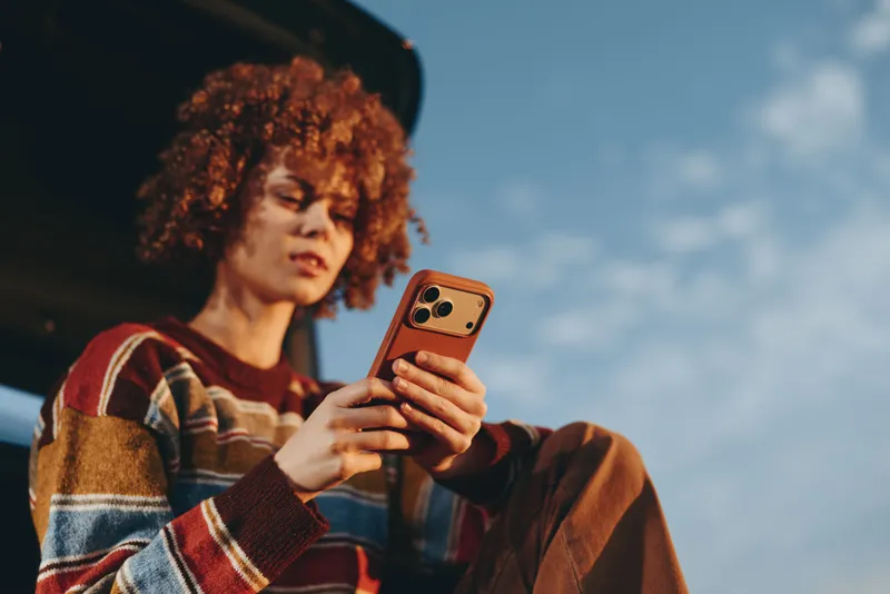 Woman with curly hair holding smartphone outdoors, casual lifestyle, blue sky background, seated and relaxed, soft daylight, communication moment, technology for everyday, life