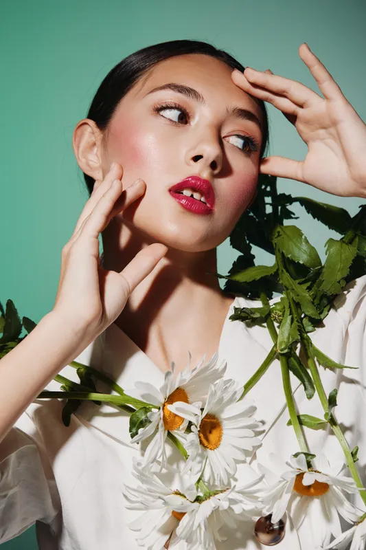 Elegant woman with flawless glossy skin, artistic makeup, and pink lipstick poses with daisies against green background. Beauty creativity and fashion concept.