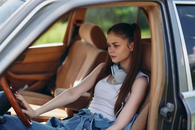 Young woman with long hair in casual attire sitting in a car, wearing headphones, enjoying a moment of relaxation in nature, conveying tranquility and leisure.