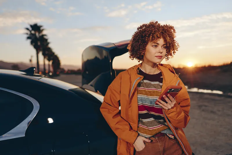 Woman lifestyle on road at sunset near car, curly hair, orange jacket, holding tablet, calm smile, evening light, travel portrait, parked vehicle background