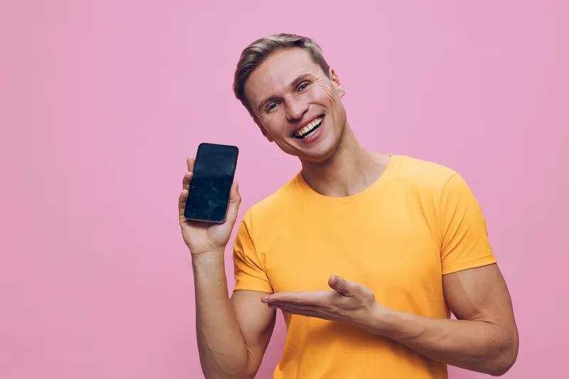 smiling man holding smartphone in yellow t-shirt showing screen isolated on pink background digital detox mental health concept