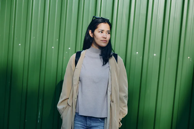 Woman in a beige coat and light gray sweater stands before a vivid green wall, showcasing casual street style and calm mood in soft daylight, urban simplicity and relaxed wardrobe statement
