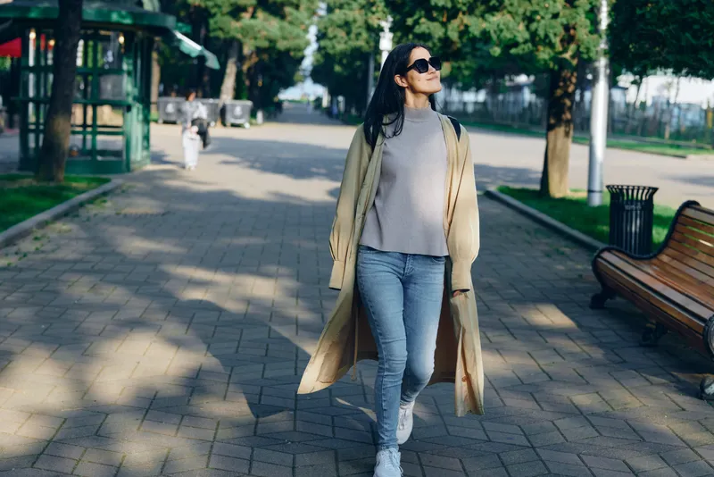 Woman walking in a sunlit park wearing a long beige coat, gray sweater, blue jeans and white sneakers, carrying a tote, enjoying a casual city stroll along a tree lined avenue in a gentle breeze