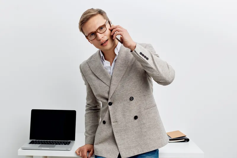 young caucasian man talking smartphone laptop on table wear grey jacket glasses business casual communication concept