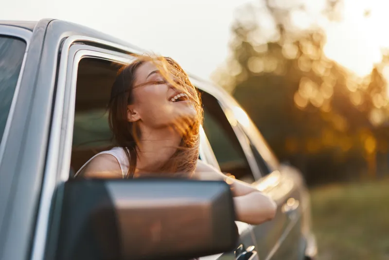 Joyful woman enjoying a sunset drive with hair flowing in the wind, exuding happiness and freedom in a car, set against a warm summer background.