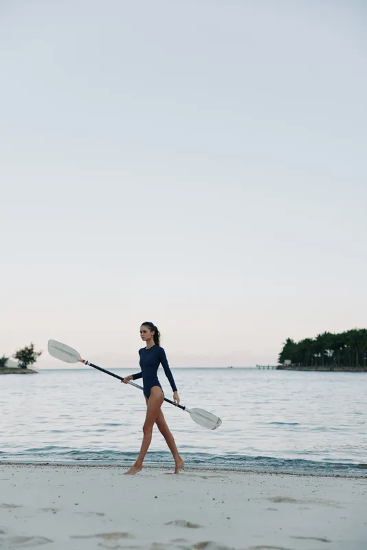 A lone figure walks along a sandy shore with two paddles, wearing a dark swimsuit as calm water and a pale sky create a serene coastal scene.