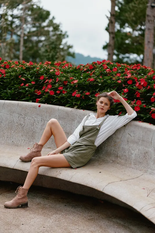 Fashionable young woman in olive green overalls and white shirt, sitting casually in a garden with vibrant red flowers, showcasing a relaxed, summer vibe