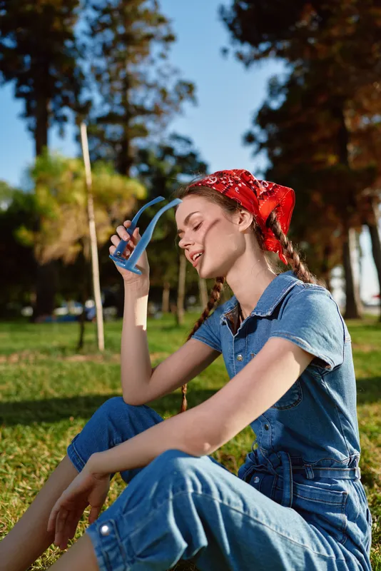 relaxed woman sitting on grass wearing denim outfit and red bandana holding sunglasses enjoying sunny day in park outdoors