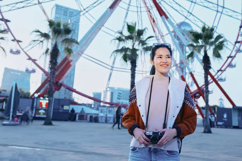Woman traveler with camera in hand smiles at the camera in an urban amusement park setting, with a large ferris wheel, palm trees and distant skyline in view