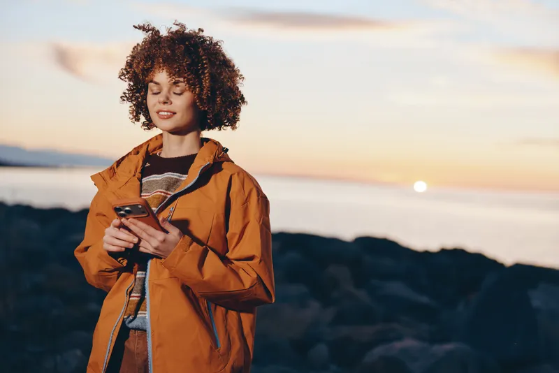 Woman lifestyle outdoors at sunset, travel jacket, curly hair, with a smile, holding travel documents, beach breeze, soft golden light, scenic coast background