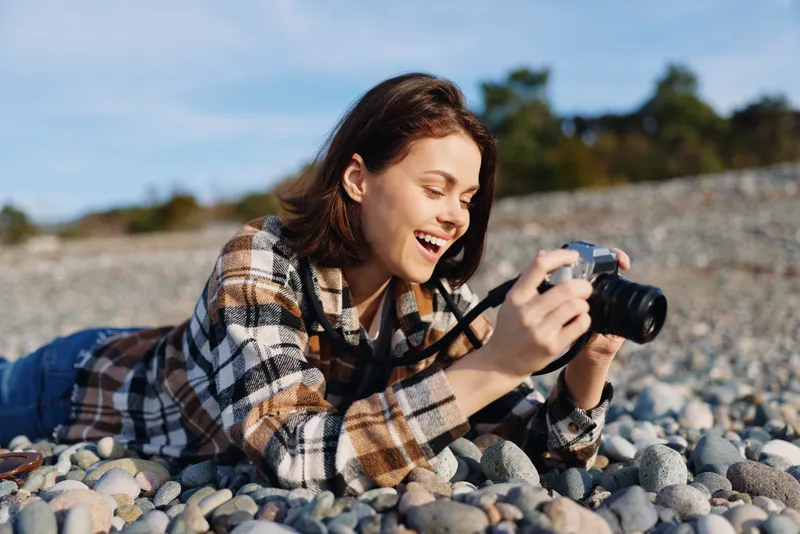 Woman photographer lying on a pebble beach, smiling while she captures candid moments with a compact camera, wearing a plaid shirt under bright sunny skies.