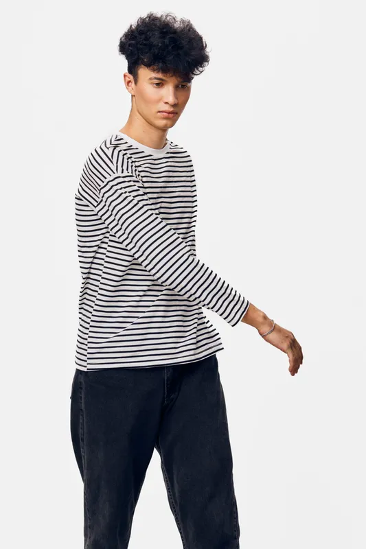 young man wearing striped long sleeve shirt and black pants standing with thoughtful expression on white background, casual style, modern look