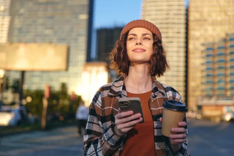 Woman with coffee and smartphone on urban street at golden hour, candid lifestyle portrait in beanie and plaid jacket, authenticity and mindful living captured with emotional storytelling and