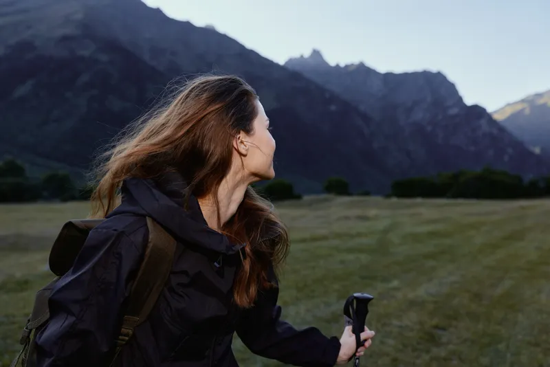 A focused hiker with a backpack stands in an open meadow, looking toward distant rugged peaks. She holds a trekking pole, exuding curiosity and readiness for adventure.