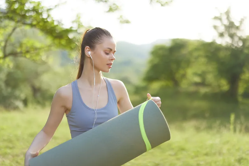 Young woman practicing yoga outdoors, holding a yoga mat, dressed in comfortable sportswear, conveying calmness and focus amidst lush greenery