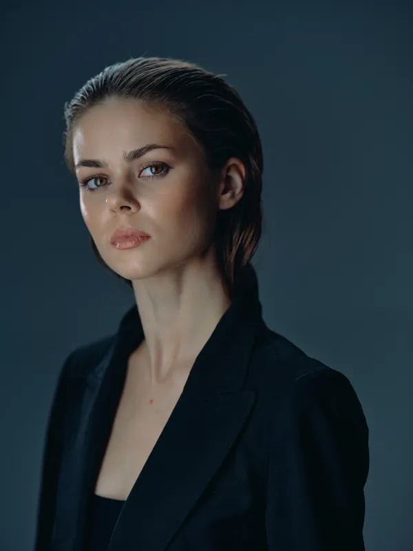 Serious woman in black jacket with slicked-back hair against dark background