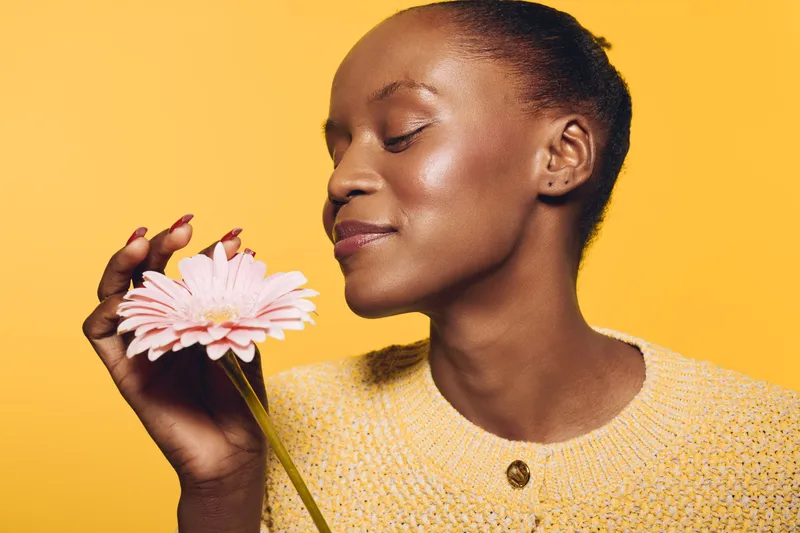 Young black woman with dark skin wearing yellow sweater gently holds pink flower close to face, eyes closed and smiling softly. Concept of natural beauty, relaxation, and floral wellness.