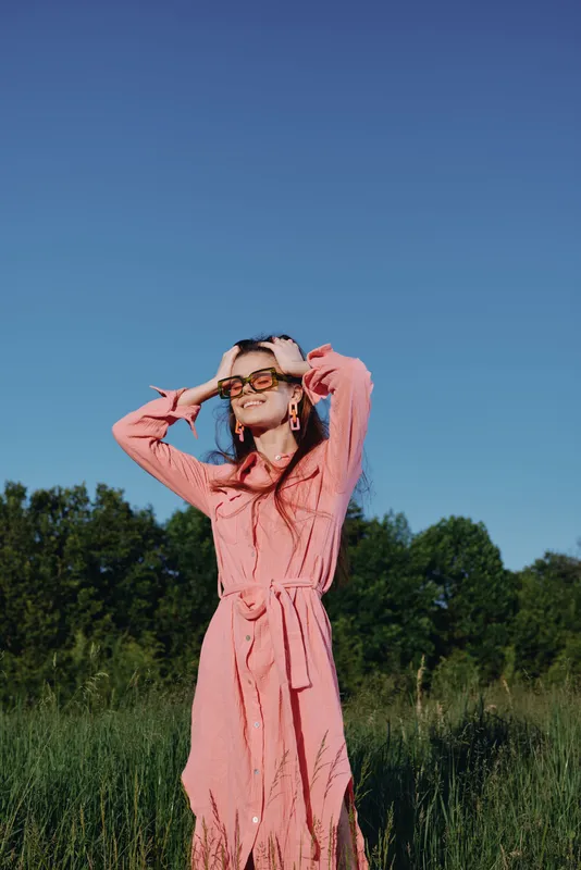 Young woman in pink dress enjoying sunny day outdoors, posing with confidence amid green nature, expressing joy and freedom in a vibrant seasonal setting
