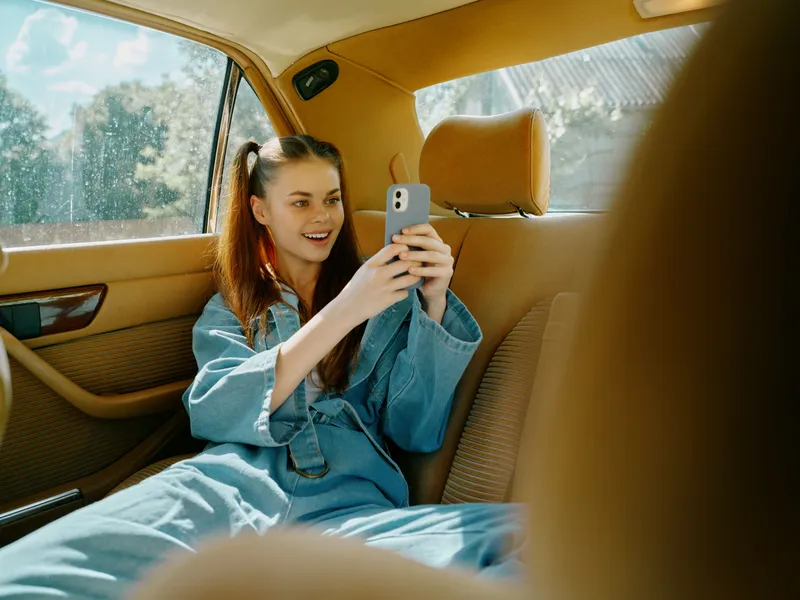 Young woman in denim clothing happily using smartphone in car, showcasing relaxed emotions and bright, cheerful atmosphere.