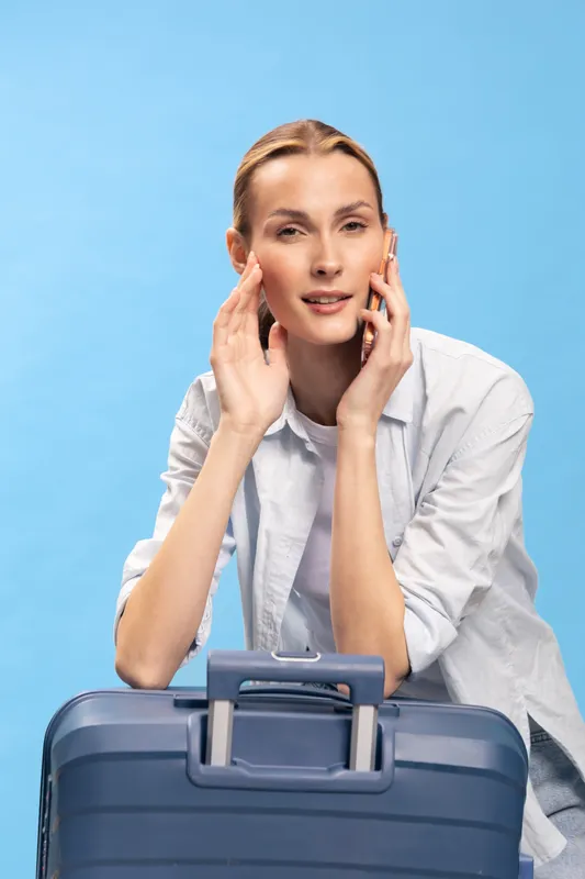 Young woman with suitcase talking on smartphone isolated on blue background. Casual travel concept with female traveler preparing for trip or vacation.