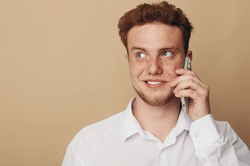 Young man with red hair and freckles wearing white shirt talks on smartphone looking away. Isolated on beige background. Concept of communication, technology, and casual lifestyle.