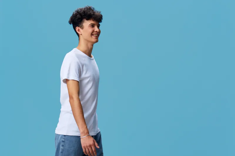 smiling man wearing white t-shirt and blue jeans stands against blue background with casual pose and curly hair, looking sideways with happy expression