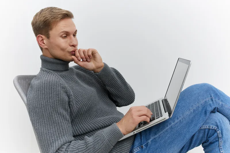thoughtful man using laptop gray sweater casual jeans sitting chair digital nomad remote work concept technology in white studio background