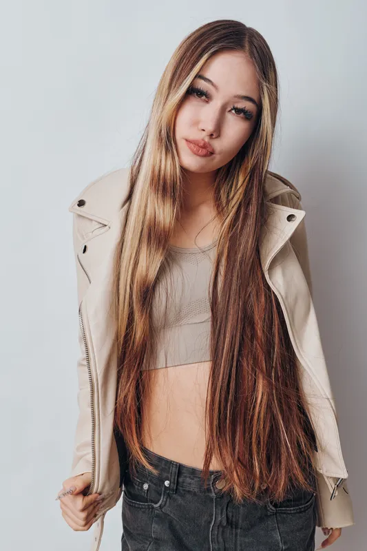 Young woman with long straight hair wearing beige cropped top and jacket posing in studio with neutral expression on white background. Casual style and fashion portrait.