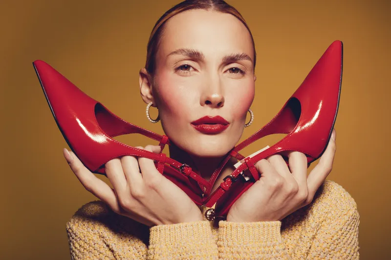 Stylish woman with red lipstick holds red high heels near face against yellow background. Fashion concept with elegant makeup, accessories, and confident female model portrait in studio lighting.