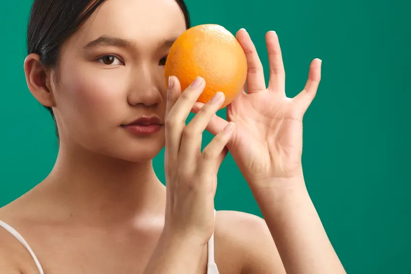 asian woman holds orange near her face against a vibrant green backdrop, capturing a calm yet artistic moment with a soft gaze and balanced composition.