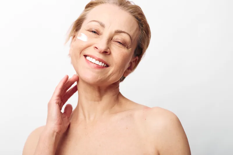 Smiling mature woman with natural skin applies white face cream on cheek, posing against white background. Concept of skincare, healthy aging, self-care, beauty routine, and wellness.
