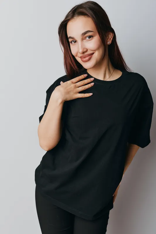 Portrait of smiling woman with dark hair wearing black t shirt standing against white background with hand on chest, casual style and confident expression in studio lighting.