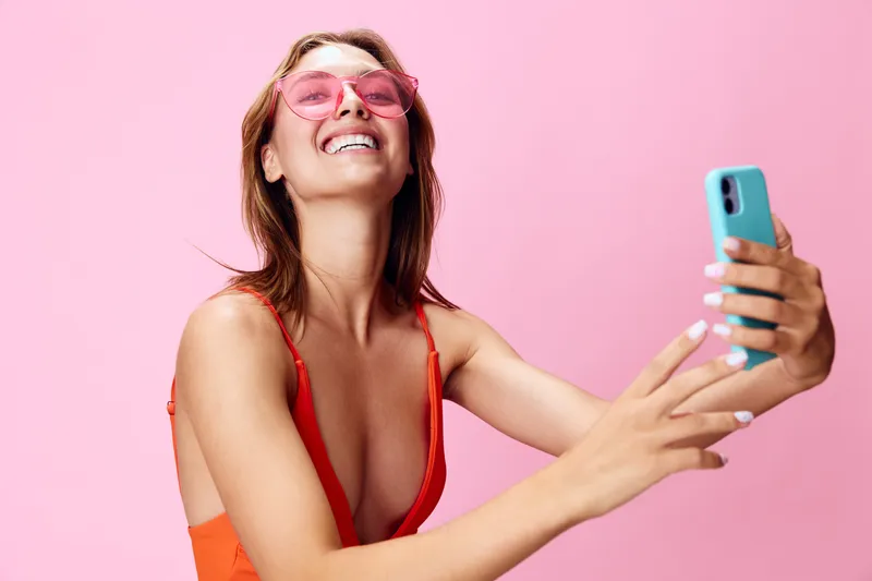 Радостная женщина в модном купальнике делает селфи на смартфон на игривом розовом фоне