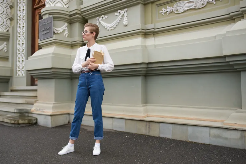 Симпатичная студентка с книгой в руке, в очках, на фоне здания в ретро-стиле — образовательное учреждение
