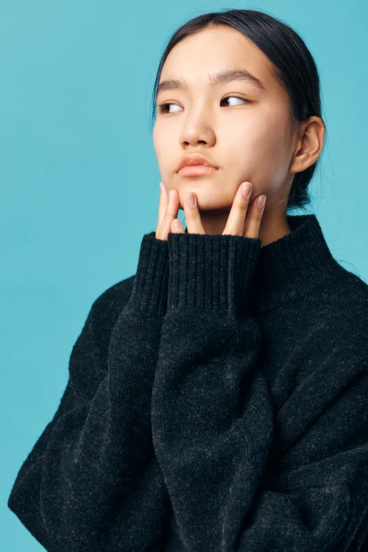 Young woman in black sweater looking away with thoughtful expression on blue background. Closeup portrait of serious female with natural makeup and long dark hair in casual outfit.