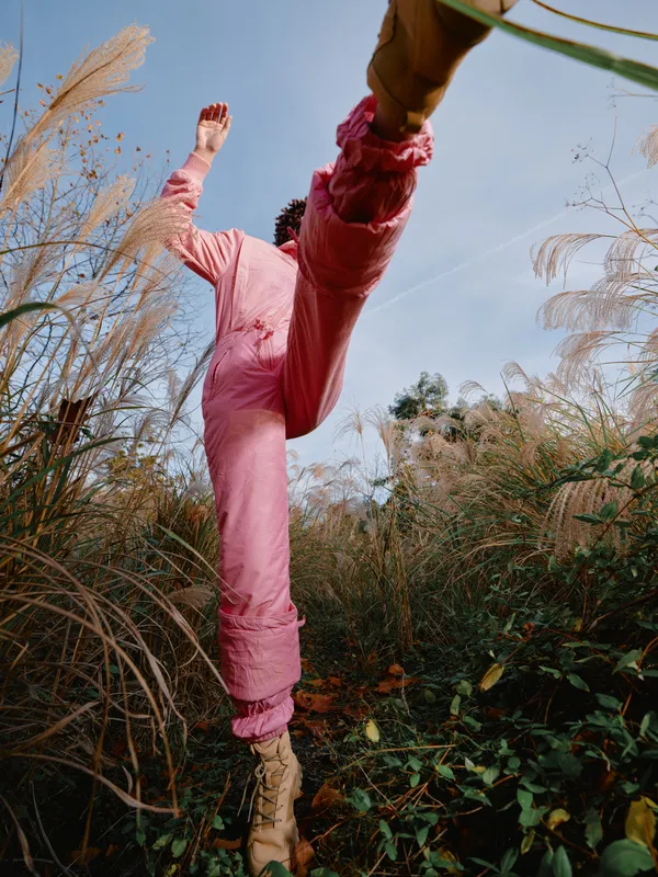 Woman jumping outdoors in pink pants among tall grasses with a blue sky, capturing energetic motion, playful nature, and vibrant spring vibes in candid active photography.