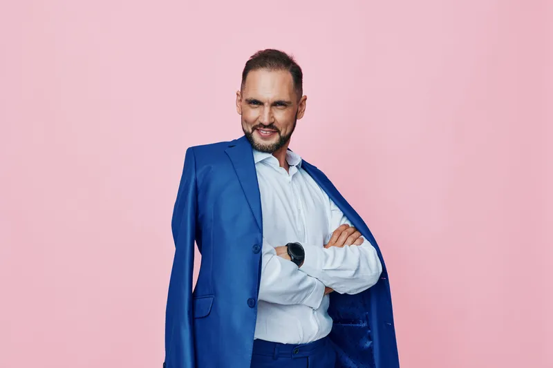 A confident business professional stands against a solid pink background, wearing a blue suit and light shirt. He projects calm confidence and poised demeanor.