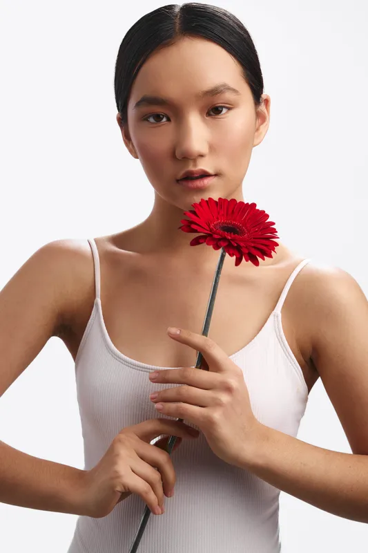 Asian woman poses with a red flower in a minimal studio setting. She gazes softly at the camera, exuding calm confidence and subtle elegance through gentle lighting.