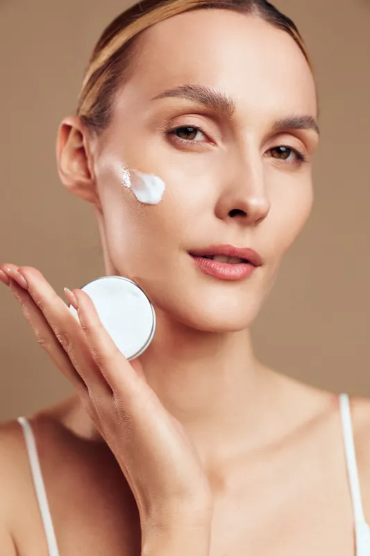 Confident woman with smooth skin applying cream to her cheek, close up portrait, soft lighting, neutral tones, skincare routine moment, healthy glow, gentle touch, natural look