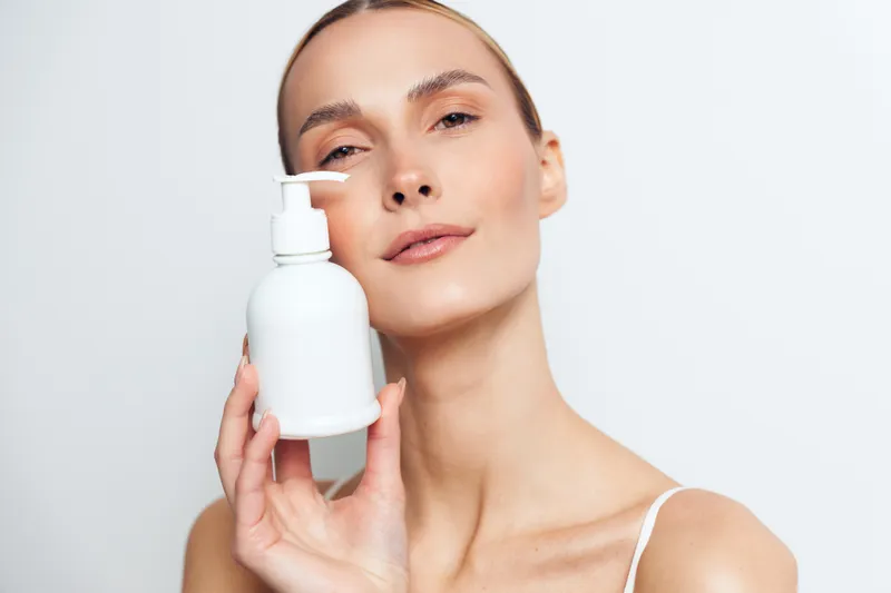 Smiling woman holding a moisturizer bottle near her face in a clean white spa setting skincare concept for hydration glow and self care beauty routine for healthy skin