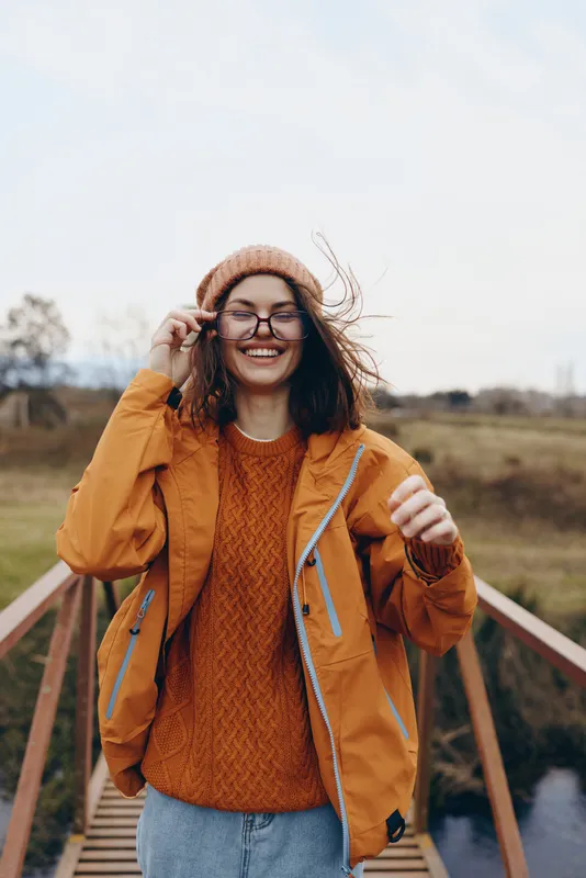 Woman outdoors lifestyle laughing hat jacket winter countryside tracks train, smiling with wind in hair, holding backpack strap, orange sweater, casual travel moment, natural colors, rural landscape