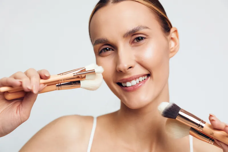 Young woman with blonde hair smiles while holding makeup brushes near her face against a plain light background in a beauty and skincare concept.