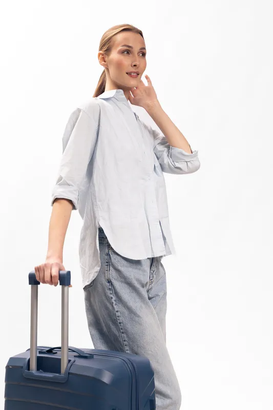 Young woman in casual clothes holding suitcase handle, looking away thoughtfully. Travel concept with luggage, preparation, and journey anticipation in bright studio setting.