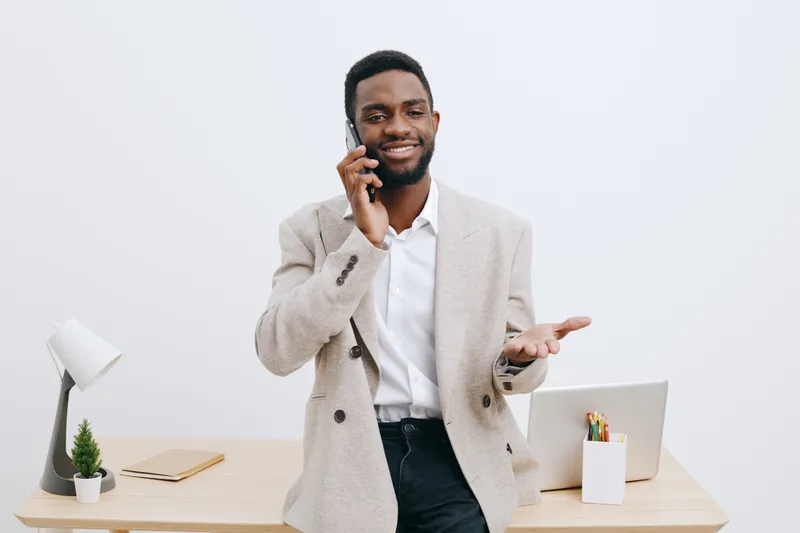 Confident smiling man with dark skin talking on mobile phone while standing at modern office desk, wearing light beige blazer and white shirt. Professional, business, communication concept with worksp