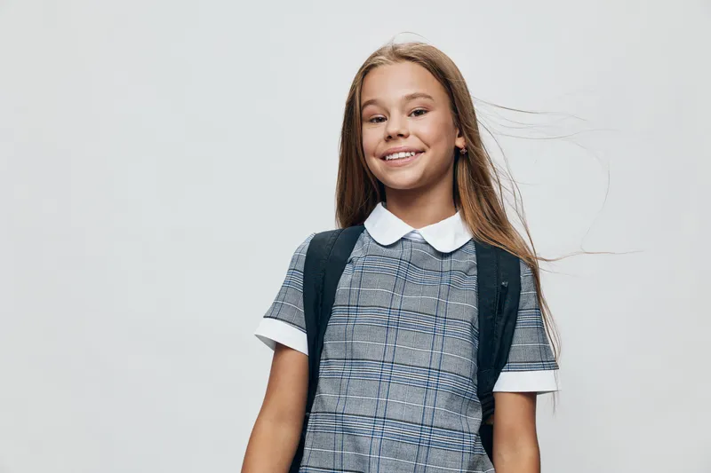 Girl smiling and standing with school backpack wearing casual plaid dress with collar and white short sleeves against light background in studio portrait.