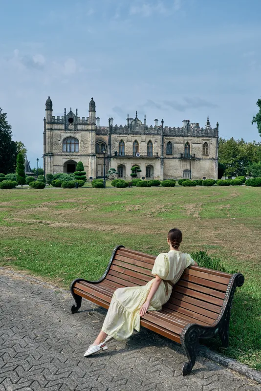 A woman in a light dress sits on a wooden bench in a park with a grand historic castle in the background creating a serene outdoor scene
