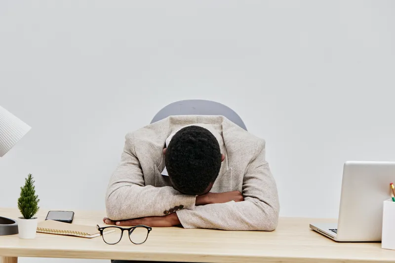 Business man exhausted at office desk, resting head on arms with closed eyes, workspace with laptop, glasses, notepad, and phone, minimalistic white background, stress and fatigue concept.