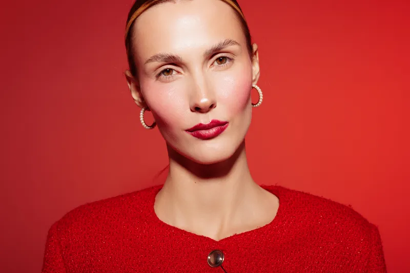 Portrait of confident woman with red lipstick and earrings wearing red sweater against red background. Studio lighting highlights smooth skin and elegant facial expression.